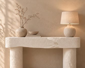 May include: A beige stone console table with a rough, textured surface and two cylindrical legs. On top sits a round vase with thin branches, a small bowl on two stacked books, and a table lamp with a textured base and a fabric shade.