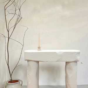 May include: White stone console table with a rough, textured surface. The table has two thick, rounded legs and a rectangular top. A small white candle holder with a single white candle is on the table.