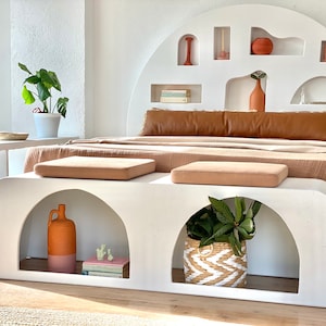 May include: A white bed frame with a curved headboard featuring multiple niches and shelves. The headboard is decorated with terracotta pots, vases, and books. The bed has a brown leather headboard cushion and a beige blanket. A white bench with two cushions sits in front of the bed. The bench has two arched niches, one with a terracotta vase and books, the other with a potted plant.
