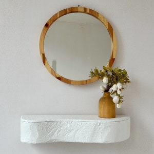 May include: A white, curved shelf with a wooden round mirror and a wooden vase with white flowers. The mirror has a natural wood frame.
