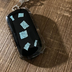 May include: A black glass pendant necklace with a silver chain. The pendant has a rectangular shape and features small, iridescent blue squares.