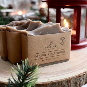 Peut inclure: Trois pains de savon rectangulaires à la surface lisse brun clair, chacun enveloppé dans une bande de papier beige étiquetée CHOICE Naturseifen Orange & Patchouli, natural soap, vegan, avec un petit logo circulaire et une icône dessinée.