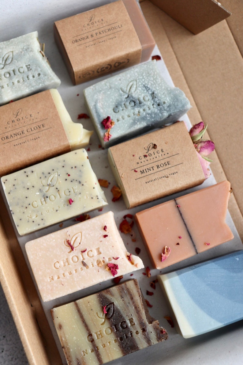 May include: A collection of eight natural soap bars in various colors and scents, including orange & patchouli, orange clove, mint rose, and more. Each bar is wrapped in brown paper with the brand name "Choice Naturals" printed on it.