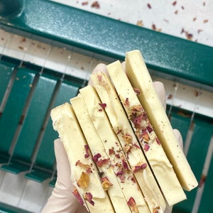 May include: A stack of five white soap bars with dried rose petals embedded in the soap. The soap bars are arranged in a hand wearing a white glove.