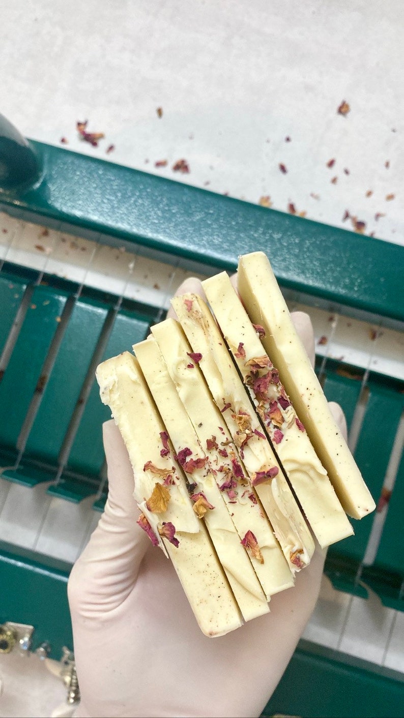 May include: A stack of five white soap bars with dried rose petals embedded in the soap. The soap bars are arranged in a hand wearing a white glove.