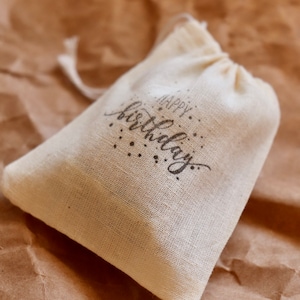 May include: A small, off-white, woven fabric drawstring pouch featuring the text "Happy Birthday" printed in a dark, stylized script font on the front, accompanied by small decorative dots surrounding the lettering.