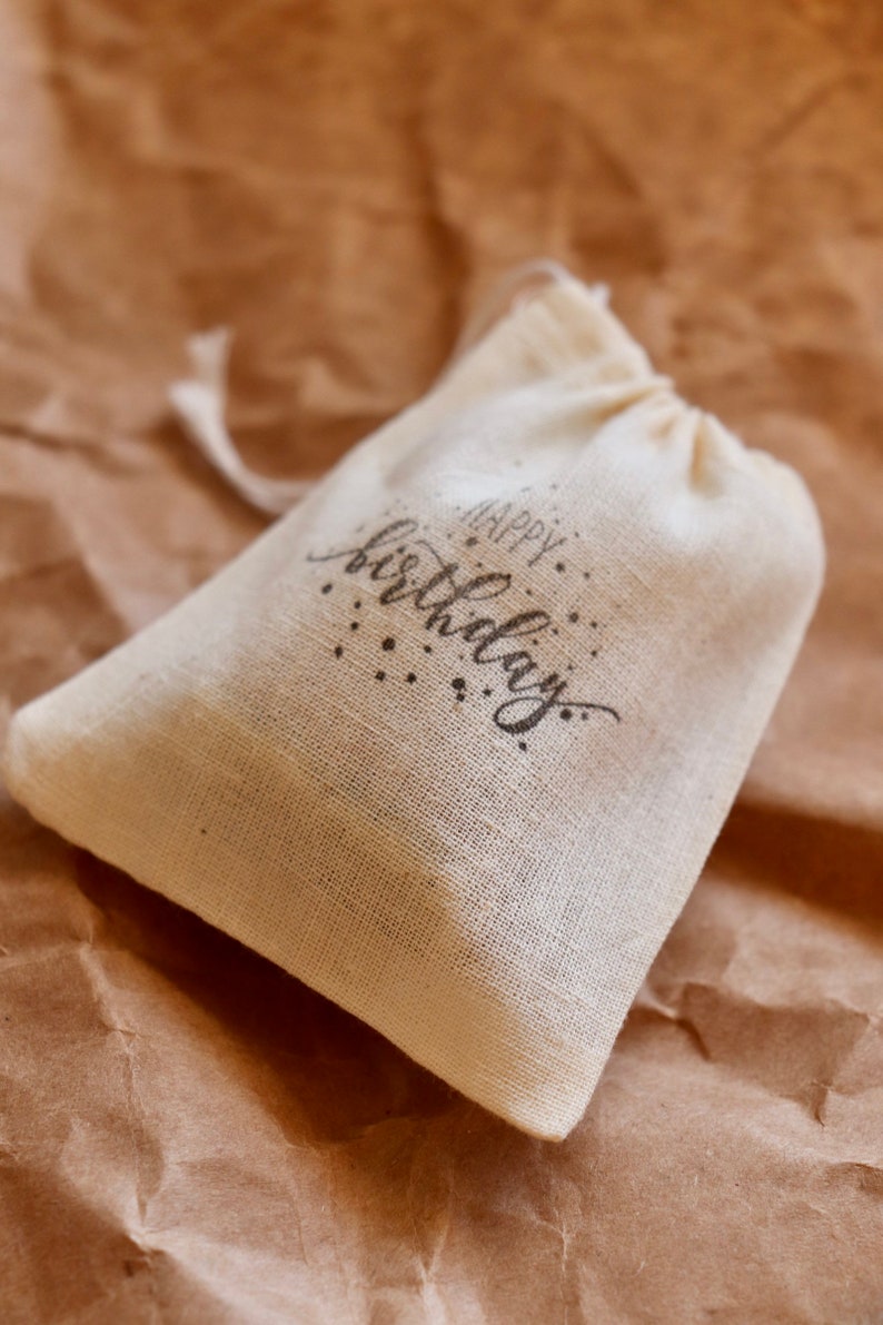 May include: A small, off-white, woven fabric drawstring pouch featuring the text "Happy Birthday" printed in a dark, stylized script font on the front, accompanied by small decorative dots surrounding the lettering.