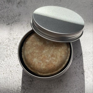 Könnte beinhalten: Eine runde, hellbraune Seife in einer silbernen Dose mit Deckel. Die Seife befindet sich in einem kleinen, runden Behälter.