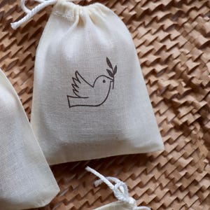 May include: A small off-white cotton drawstring pouch featuring a dark brown line art illustration of a bird holding an olive branch in its beak. The bag is made of a woven fabric and includes a white cord closure at the top for securing contents.