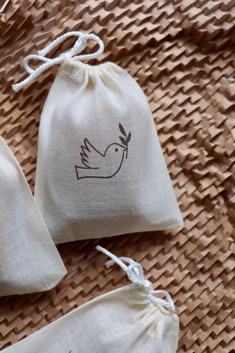 May include: A small off-white cotton drawstring pouch featuring a dark brown line art illustration of a bird holding an olive branch in its beak. The bag is made of a woven fabric and includes a white cord closure at the top for securing contents.