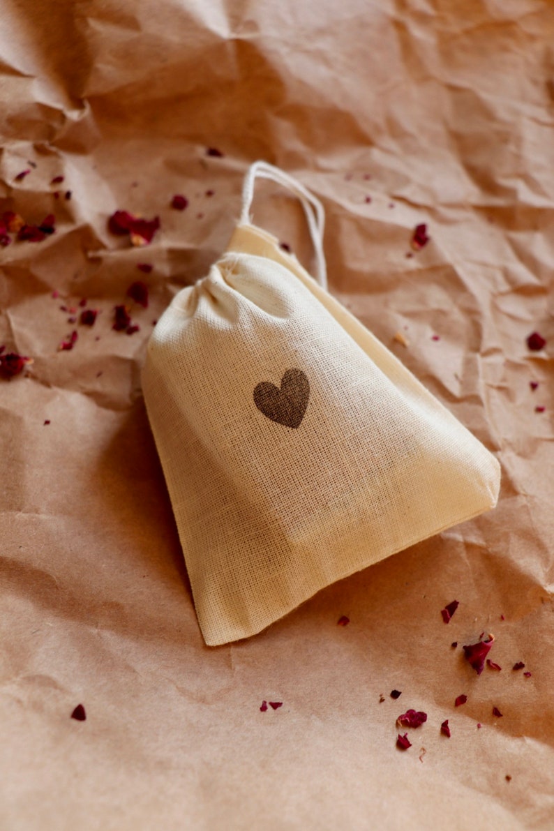 May include: A small, off-white fabric drawstring pouch featuring a simple, dark brown heart icon printed on the front. The bag is made of a woven textile material and includes a thin white cord at the top for closure, creating a gathered effect.