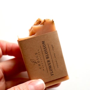 May include: A rectangular bar of peach-colored soap topped with dried flower petals. The soap is wrapped in a brown paper label printed with the text "CHOICE NATURSEIFEN FLOWER BLOSSOM natural | soap | vegan" and a small leaf icon.