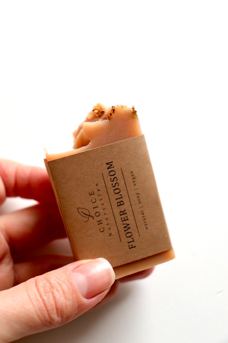 May include: A rectangular bar of peach-colored soap topped with dried flower petals. The soap is wrapped in a brown paper label printed with the text "CHOICE NATURSEIFEN FLOWER BLOSSOM natural | soap | vegan" and a small leaf icon.