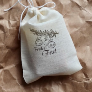 May include: A small off-white fabric drawstring pouch featuring a dark brown printed graphic of a pine branch with three hanging ornaments and the text Frohes Fest. The bag is made of a woven textile material and is closed with a white string.