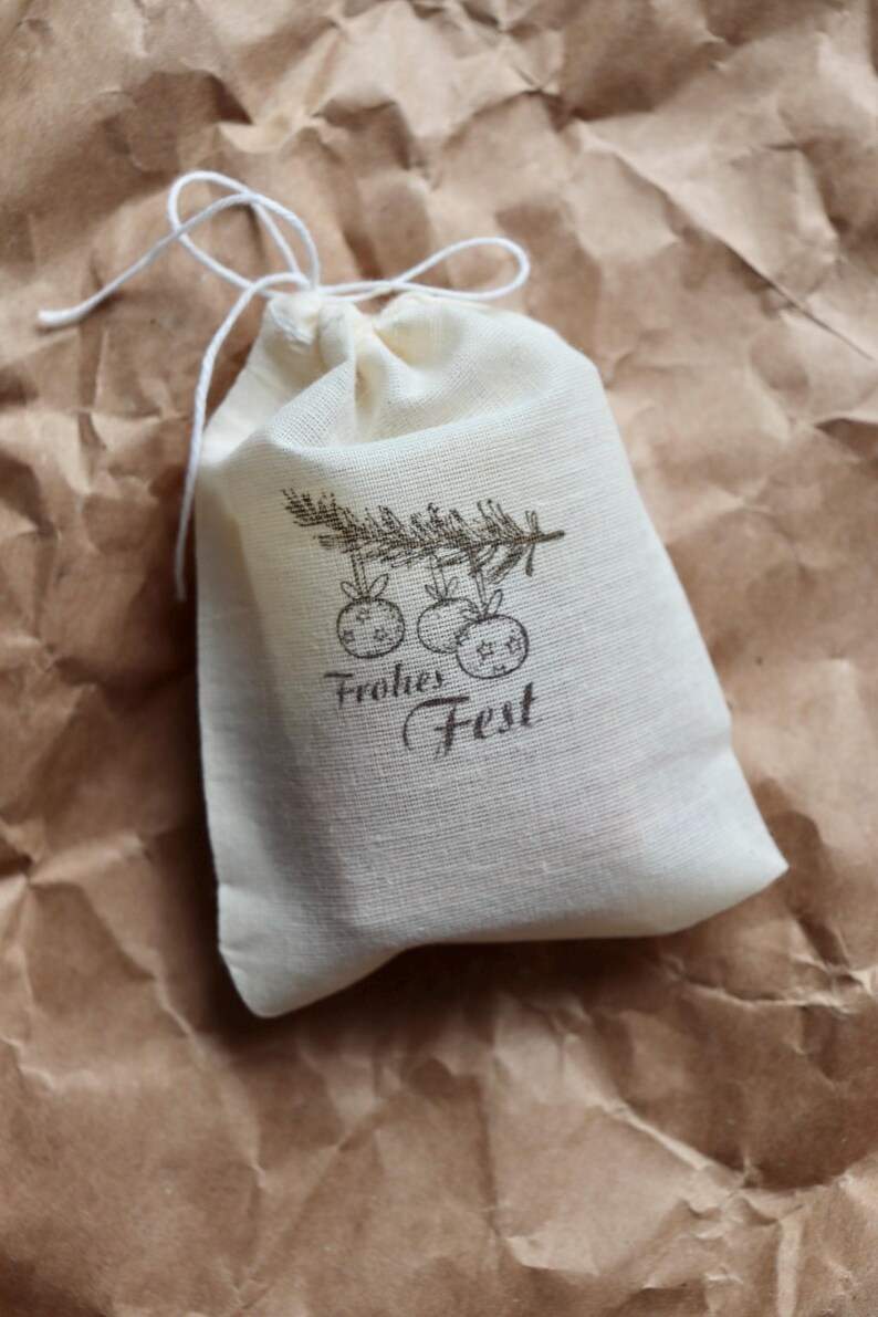 May include: A small off-white fabric drawstring pouch featuring a dark brown printed graphic of a pine branch with three hanging ornaments and the text Frohes Fest. The bag is made of a woven textile material and is closed with a white string.