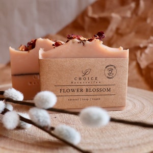 May include: Two rectangular bars of light pink soap topped with dried flower petals. Each bar is wrapped in a brown paper label featuring the text CHOICE Naturseifen, FLOWER BLOSSOM, and natural | soap | vegan, with a circular handcrafted product logo.