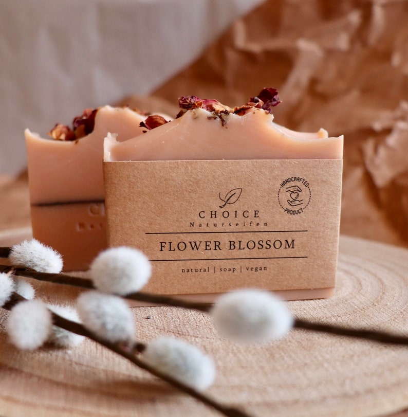 May include: Two rectangular bars of light pink soap topped with dried flower petals. Each bar is wrapped in a brown paper label featuring the text CHOICE Naturseifen, FLOWER BLOSSOM, and natural | soap | vegan, with a circular handcrafted product logo.