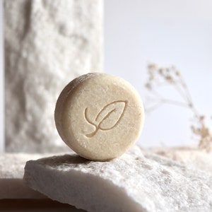 Könnte beinhalten: Ein runder, weißer Shampoo-Bar mit einem geprägten Blattdesign auf der Oberseite. Der Bar liegt auf einer weißen, strukturierten Oberfläche.