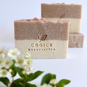 Puede incluir: Tres barras de jabón natural de color marrón claro y blanco. El jabón está cubierto con cristales de sal rosa del Himalaya. El jabón está estampado con las palabras "CHOICE Naturseifen".