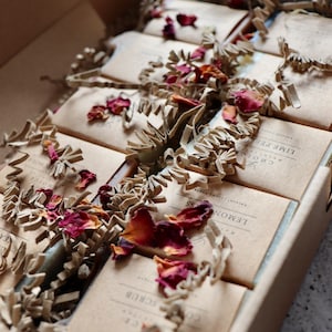 May include: A box filled with handmade soap bars wrapped in brown paper. The soaps are arranged in rows and are surrounded by shredded paper and dried rose petals. The soap labels include the names "Lemon Grass", "Lime Peel", and "Coffee Scrub".