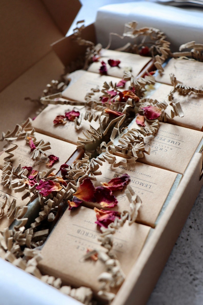 May include: A box filled with handmade soap bars wrapped in brown paper. The soaps are arranged in rows and are surrounded by shredded paper and dried rose petals. The soap labels include the names "Lemon Grass", "Lime Peel", and "Coffee Scrub".