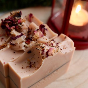 May include: Two rectangular bars of light beige soap topped with dried rose petals and botanical fragments. The soap has a smooth texture with a thin dark horizontal line running through the center of each bar, creating a layered appearance.
