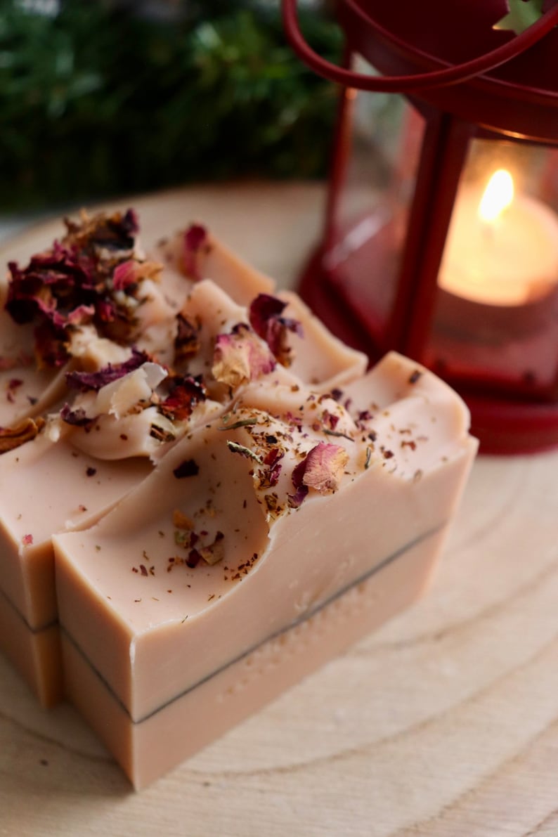 May include: Two rectangular bars of light beige soap topped with dried rose petals and botanical fragments. The soap has a smooth texture with a thin dark horizontal line running through the center of each bar, creating a layered appearance.