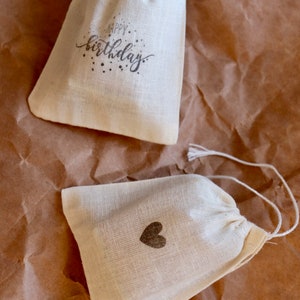 May include: Two off-white fabric drawstring pouches. One pouch features the text Happy Birthday in black script with small dots. The other pouch displays a small, solid black heart icon centered on the front. Both bags have white string ties.