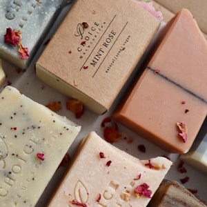 May include: Assortment of natural soap bars with the brand name "Choice Naturseifen" printed on each bar. The bars are in various colors and textures, including a light gray bar with a dark gray swirl, a light brown bar with a dark brown swirl, a light beige bar with a dark brown swirl, and a light pink bar with a dark brown swirl. The bars are arranged on a white surface with dried rose petals scattered around them.
