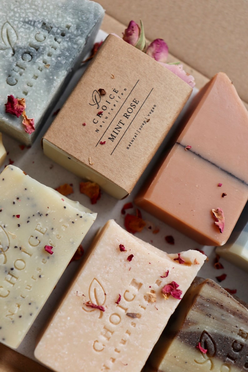 May include: Assortment of natural soap bars with the brand name "Choice Naturseifen" printed on each bar. The bars are in various colors and textures, including a light gray bar with a dark gray swirl, a light brown bar with a dark brown swirl, a light beige bar with a dark brown swirl, and a light pink bar with a dark brown swirl. The bars are arranged on a white surface with dried rose petals scattered around them.