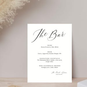 Puede incluir: Un cartel blanco con texto negro que dice "The Bar" y enumera las opciones de bebidas disponibles: Vino, Cerveza, Cócteles de Autor y Bebidas Sin Alcohol. El cóctel de autor es "The Newlywed Mule" con vodka, jugo de lima y cerveza de jengibre. Las bebidas sin alcohol son jugo de naranja, soda y agua. El cartel está firmado "The Bride & Groom" con la fecha 23 de enero de 2023.