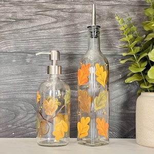May include: A clear glass soap dispenser with a silver pump and a clear glass oil bottle with a silver pour spout, both decorated with hand-painted orange, yellow, and green oak leaves. The items feature a smooth, transparent finish.