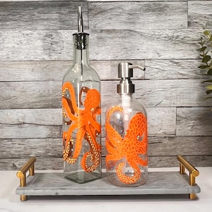 Puede incluir: Una botella dispensadora de aceite de vidrio transparente y un dispensador de jabón de vidrio transparente, ambos con diseños de pulpo naranja pintados a mano con ventosas detalladas, sobre una bandeja rectangular de mármol gris con asas doradas.