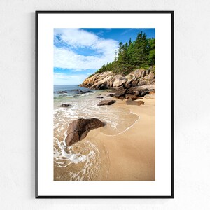 May include: Framed print of a scenic beach with a sandy shore, large rocks, and clear water. The sky is blue with white clouds, and green trees grow on a rocky cliff. The print is in a black frame.