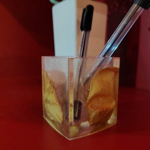 May include: A clear glass pen holder with a gold glitter resin base. Two black pens are in the holder.