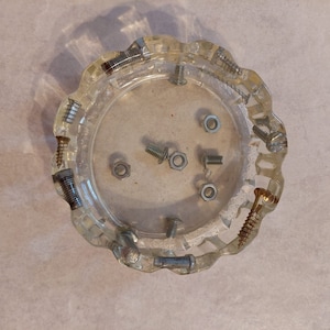 May include: A clear glass ashtray with a scalloped edge, filled with various screws and nuts. The screws are silver and gold, and the nuts are silver.