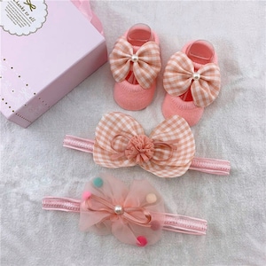 Puede incluir: Un conjunto de accesorios para bebés que incluye dos pares de calcetines rosas con lazos grandes de cuadros y detalles de perlas, y dos diademas a juego. Una diadema tiene un lazo de cuadros y la otra un lazo transparente con pompones de colores.