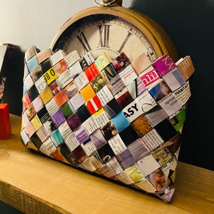 May include: A woven clutch bag made from colorful magazine pages. The bag is sitting on a wooden shelf in front of an antique clock.