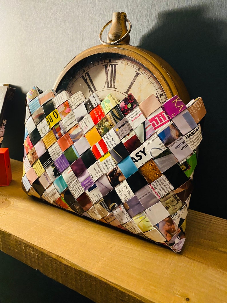 May include: A woven clutch bag made from colorful magazine pages. The bag is sitting on a wooden shelf in front of an antique clock.