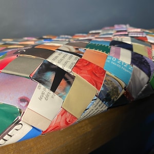 May include: A woven basket made from recycled magazine pages. The basket is woven in a diamond pattern and features a variety of colors and images. The basket is sitting on a wooden surface.
