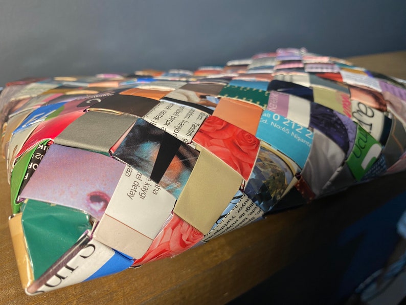 May include: A woven basket made from recycled magazine pages. The basket is woven in a diamond pattern and features a variety of colors and images. The basket is sitting on a wooden surface.