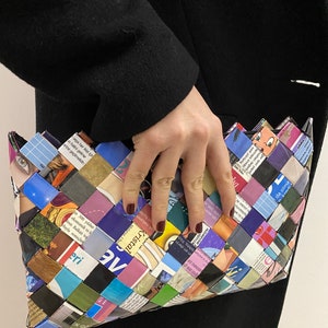 May include: A colorful clutch purse woven from recycled magazine pages. The purse is a rectangular shape with a slightly pointed top. The woven pattern is intricate and features a variety of colors, including blue, green, purple, pink, and yellow.