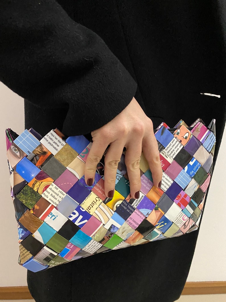May include: A colorful clutch purse woven from recycled magazine pages. The purse is a rectangular shape with a slightly pointed top. The woven pattern is intricate and features a variety of colors, including blue, green, purple, pink, and yellow.