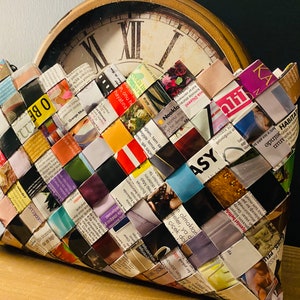 May include: A woven clutch bag made from strips of colorful magazine pages. The bag is woven in a checkered pattern and has a brown leather handle.