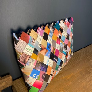 May include: A woven clutch bag made from recycled magazine pages. The bag is a patchwork of colorful magazine clippings and has a unique, geometric design.