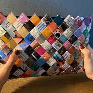 May include: A colorful woven clutch bag made from recycled magazine pages. The bag is woven in a diamond pattern and features a variety of colors and patterns.