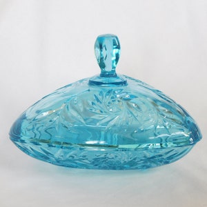May include: A blue glass candy dish with a lid and a swirl design. The dish is shaped like a heart and has a handle on the lid.