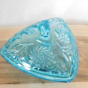 May include: A light blue glass candy dish with a triangular shape and a floral design. The dish has a lid with a knob on top.