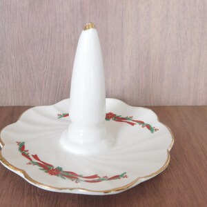 May include: White ceramic ring holder with a central cone and a scalloped base. The base is decorated with red ribbons, green leaves, and gold trim. The top of the cone has a gold accent.
