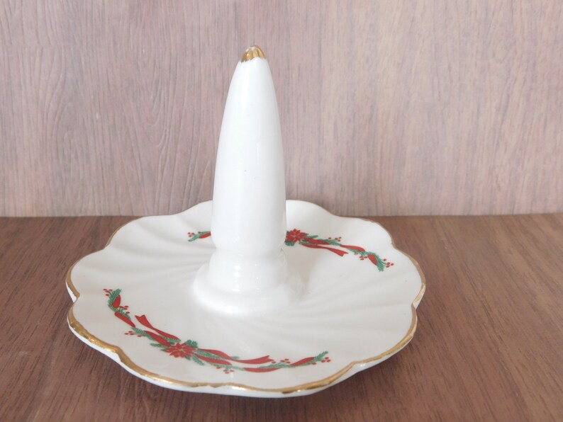 May include: White ceramic ring holder with a central cone and a scalloped base. The base is decorated with red ribbons, green leaves, and gold trim. The top of the cone has a gold accent.